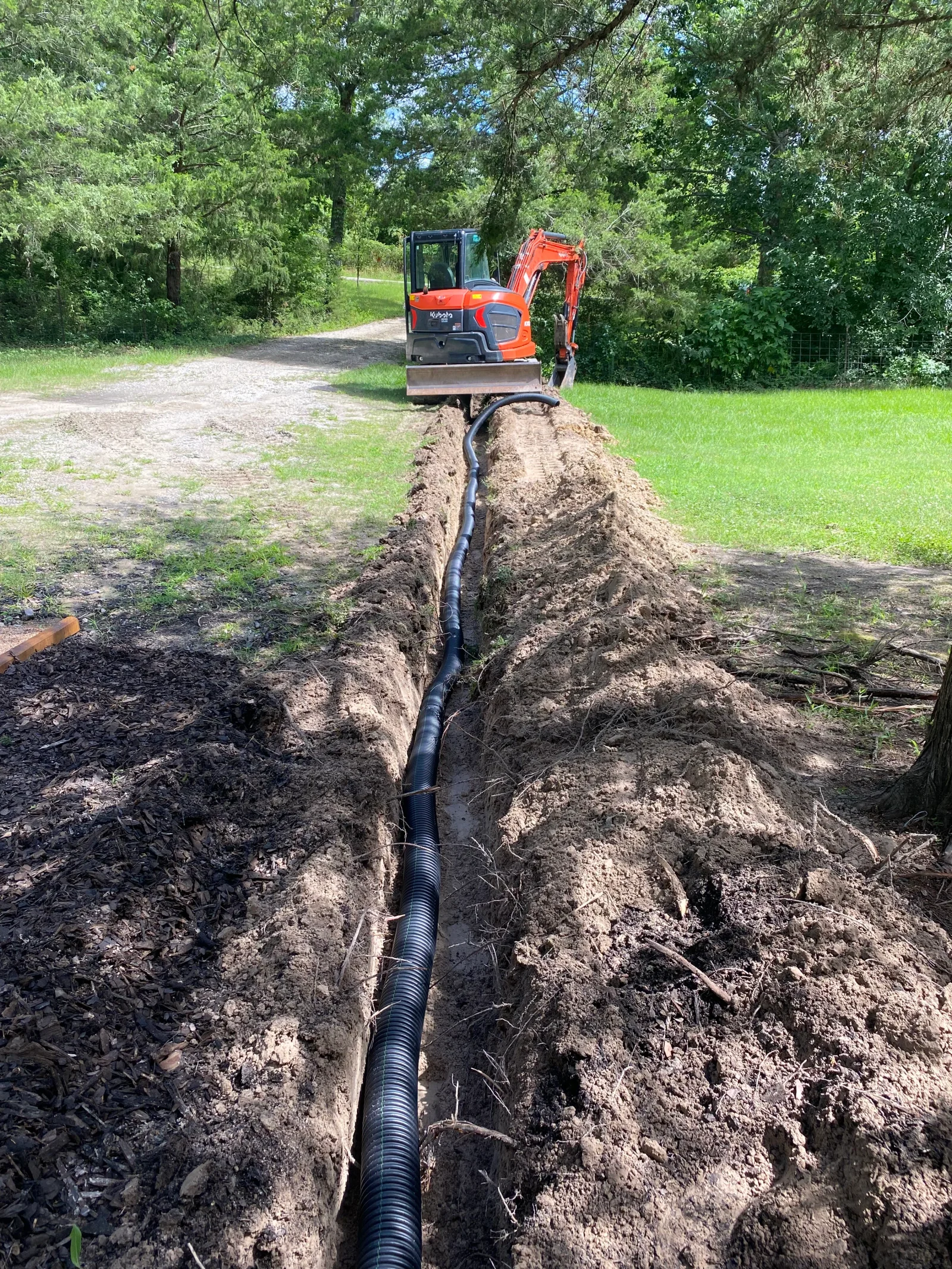 Black corrugated drainage pipe installed in excavated trench with compact excavator on site