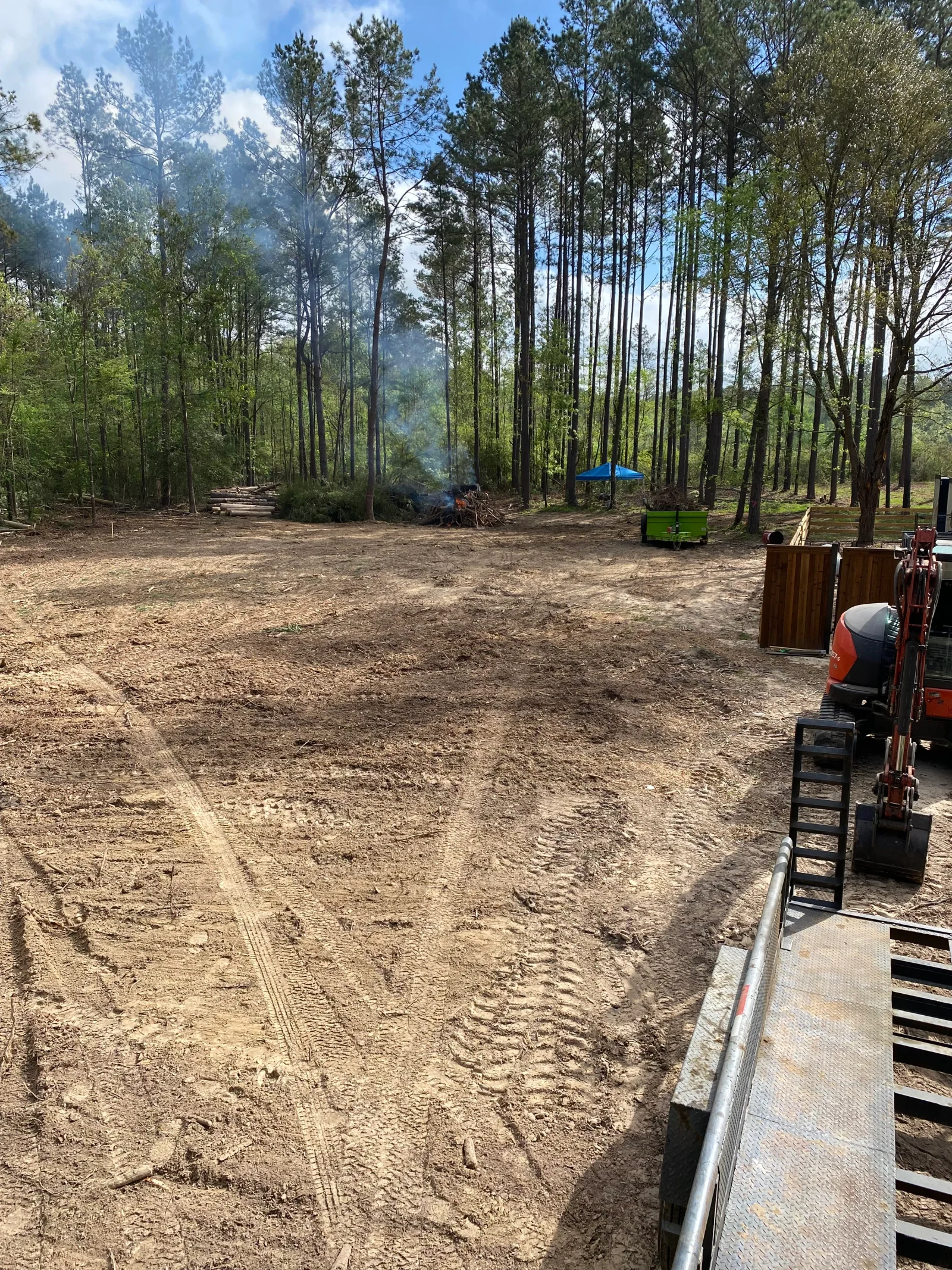 Cleared forest lot with graded dirt surface, pine trees, and equipment staging area
