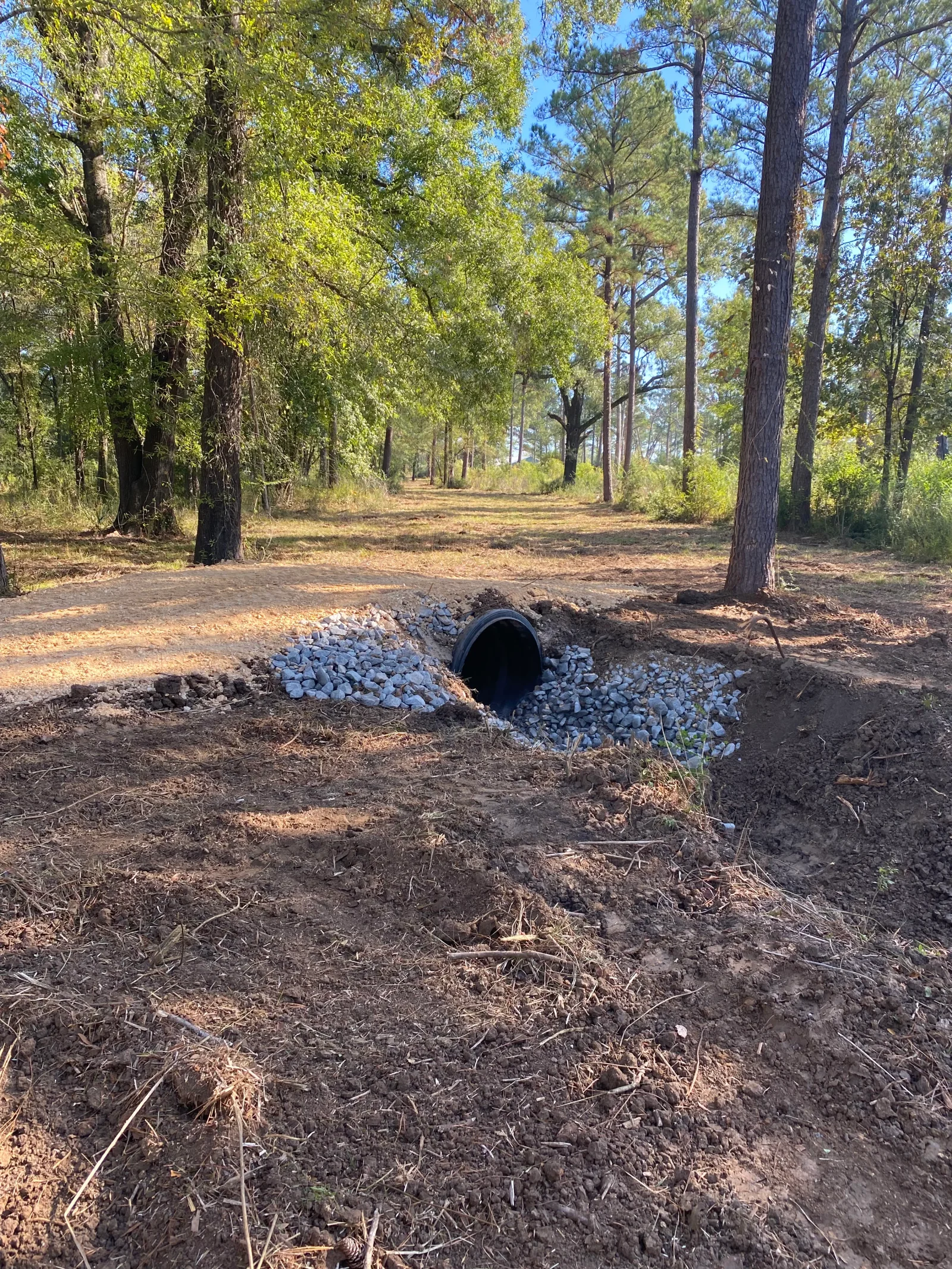 Perforated drainage pipe installed in excavated trench with crushed stone backfill along wooded driveway